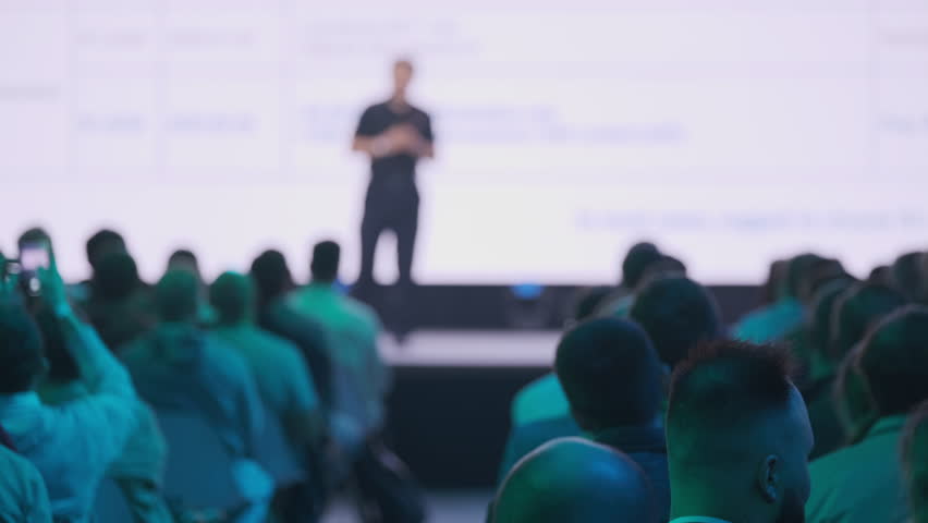 Speaker presenting on stage to a seated audience during business seminar. Conference attendees listening to informative talk.