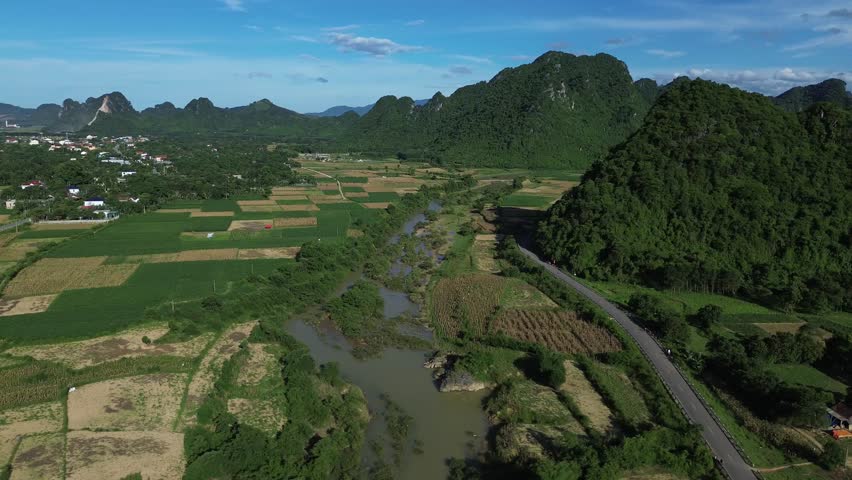Pan left aerial of rural Vietnam with winding river, cultivated fields and dense karst formations under sunny sky