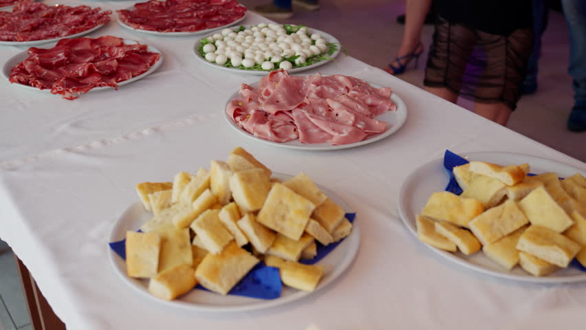 Moving shot of a party table covered with appetizers, cold cuts, cheese, and drinks, while people are celebrating nearby.