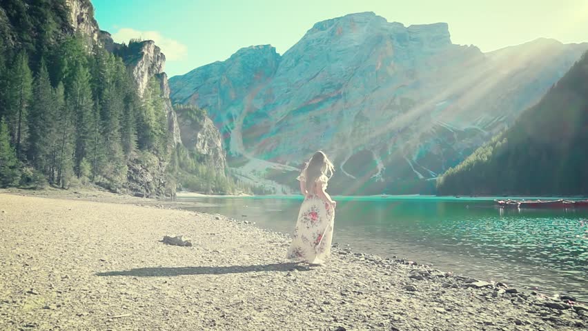 modern forest fairy on shingle birch of Lake Braies, spring light white dress with floral pattern, Spirits in water, forest and mountain landscape, girl long hair flying in wind, turned away, no face.