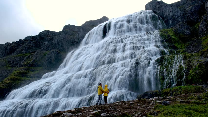 Experience the magnificence of a cascading waterfall in Iceland. A couple stands mesmerized by the stunning scenery, capturing a magical moment in a serene landscape of natures wonders.