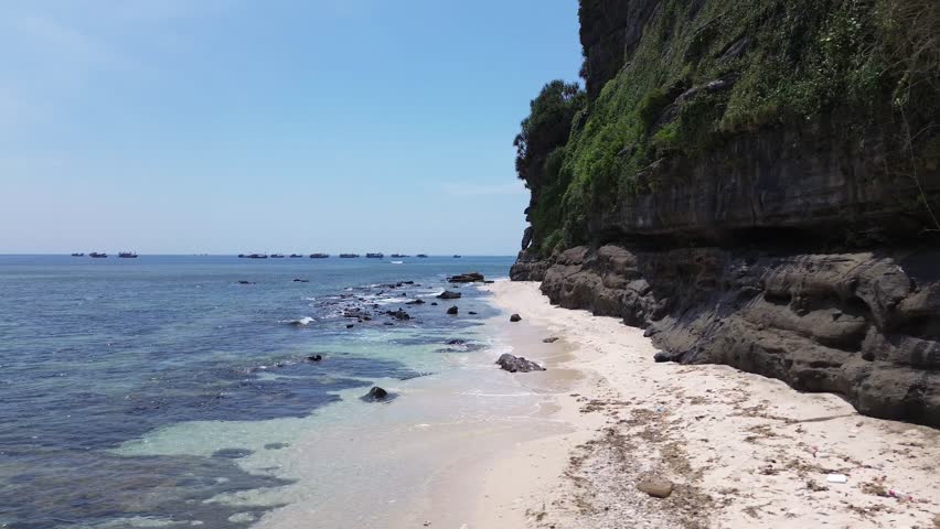 Pan left revealing a white sand beach and cliff covered in vegetation, filmed on a bright sunny day in Ly Son Island.