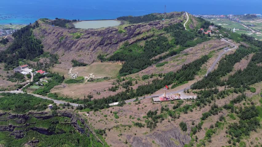 Zoom out revealing rocky volcanic cliffs, rural buildings and sea in Ly Son Island, Vietnam on a sunny afternoon.