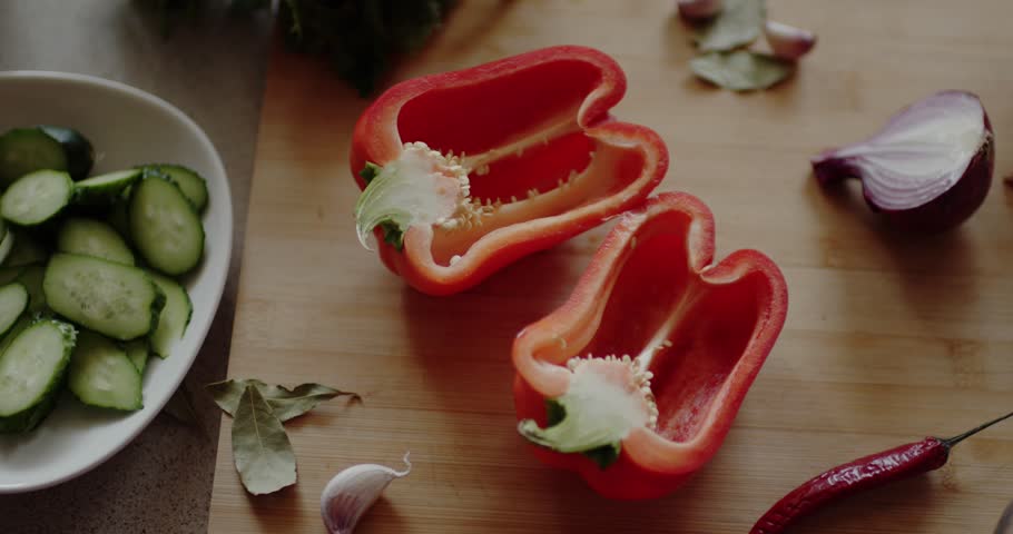 Top-down shot. A red bell pepper, sliced in half with seeds visible, is centered on a wooden board. Surrounding it are a wedge of red onion, garlic cloves, bay leaves, a chili pepper, and a plate of s