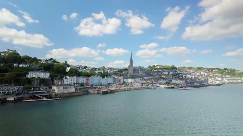 View from a moving ferry or cruise ship as it passes the picturesque town of Cobh, Ireland. The scene includes the vibrant waterfront buildings, historic St. Colman’s Cathedral, and a clear sunny sky.