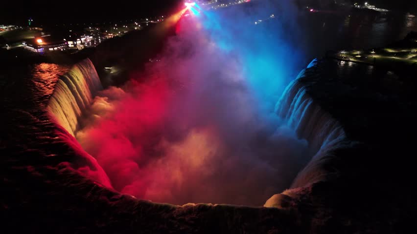 Drone circles Horseshoe Falls at night, showcasing glowing water currents while revealing busy traffic flowing below on nearby roads under the city lights