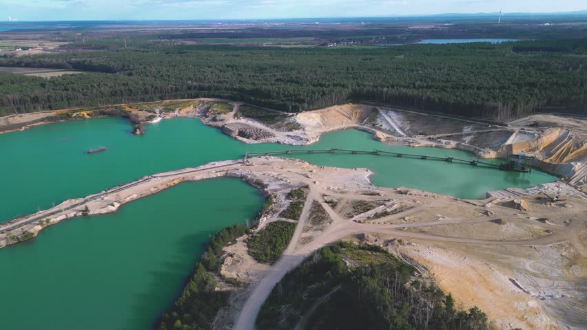 A drone shot of a sand quarry filled with turquoise water, surrounded by forest near the horizon. The open-pit mine with clear blue lakes, sand dunes, and industrial equipment