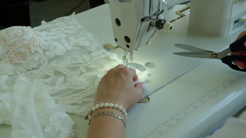 Seamstress’s hands cut thread with scissors after stitching on a sewing machine at a wedding dress production workshop