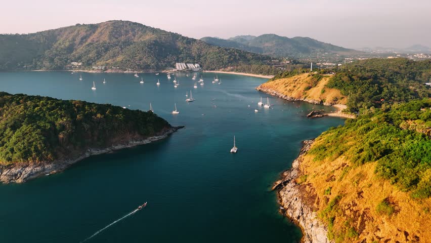 Sunset on promthep cape in Phuket, Thailand, Pristine turquoise waters surrounding white sandy coastlines and verdant hillsides framing sailing boats.