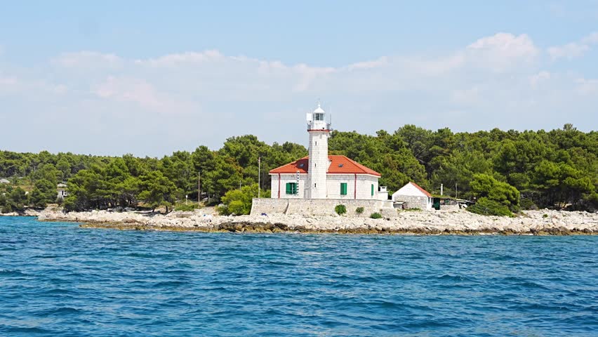 Stone lighthouse with red roof on rocky coast of Hvar Island, Croatia, surrounded by pine trees and clear blue sea. Navigation, solitude and maritime heritage in the Adriatic. View from sailing yacht