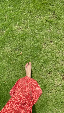 POV. First person view of barefoot run on the grass. Barefoot woman in red patterned pants running on lush green grass, top down perspective. A lively summer outdoor moment of connecting with nature.