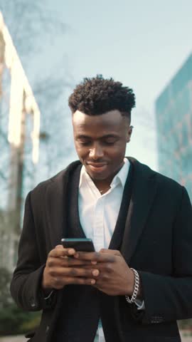 Young adult rich man walking alone in modern business district in metropolis. Medium portrait in motion of handsome black guy with modern smartphone in hand, townsman enjoying walk in center of city