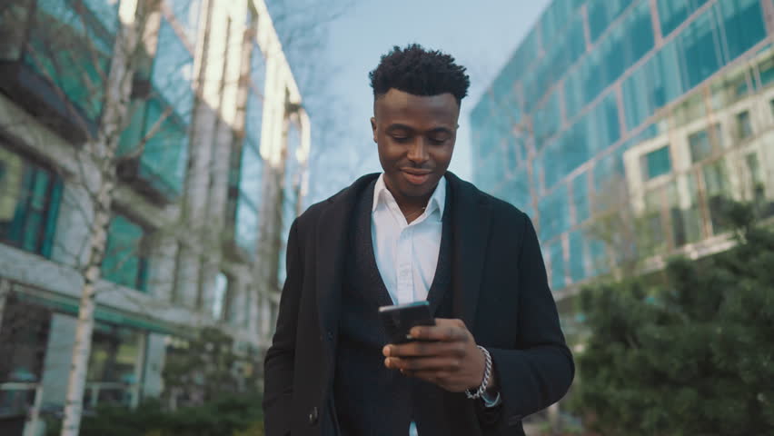 Portrait of self-confident black man walking alone in city center, good mood. Happy smiling male person strolling in downtown, successful African businessman enjoying life holding smartphone in hand