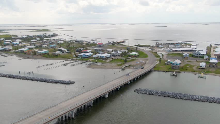 An aerial midshot of Chenier Caminada, a fishing community near Grand Isle, Louisiana. Footage includes breakwaters, coastal homes, businesses, blighted homes, vehicles, and remnants of docks.
