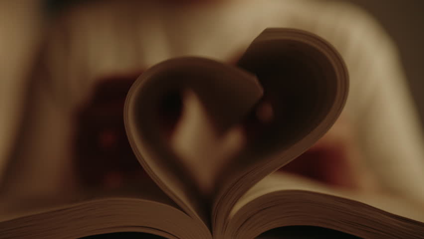 Close-up of man opening book with pages folded into heart shape, symbolizing love of reading in warm dimly lit home environment, creating cozy and inviting atmosphere. Shooting in slow motion.