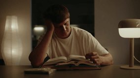 Tired male student concentrating reading two paper books sitting at a table in dark living room interior, illuminated by warm glow of modern lamps, creating studious atmosphere, close-up portrait. - Powered by Shutterstock - Get 15% off with code: PIKWIZARD15