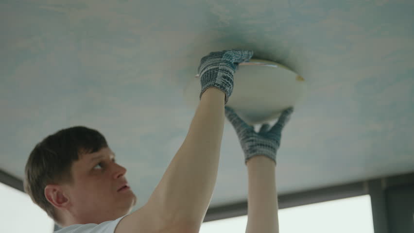 Professional electrician wearing protective gloves installing ceiling light fixture, connecting wiring and mounting electrical component in living room. Concept of electricity maintenance, slow motion