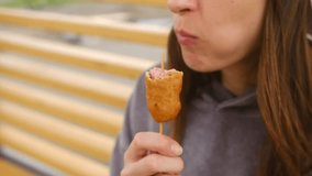 Young woman savoring corn dog, enjoying street food outdoors with cheerful expression, biting into crispy fried snack while experiencing casual dining pleasure. Woman eating corn dog on a stick - Powered by Shutterstock - Get 15% off with code: PIKWIZARD15