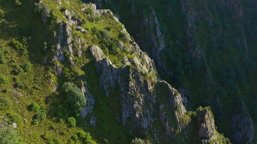 Aerial drone shot of a steep, rocky mountain cliff in Kyrgyzstan, showcasing lush greenery and dramatic terrain. The camera moves left to right, revealing depth through parallax.