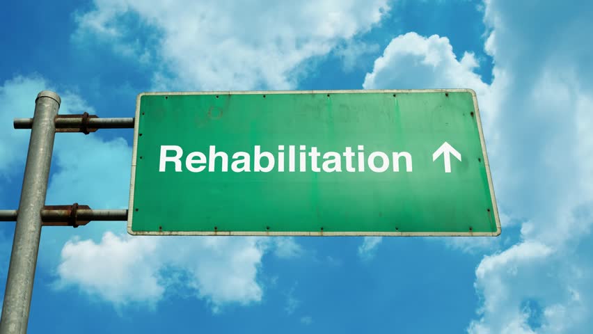 Rehabilitation text or word on a green road sign under a cloudy clean blue sky. Represents recovery, support, healing journey, and a positive path forward