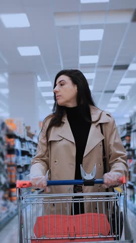 A woman, dressed in a trendy trench coat, pushes a shopping cart through a supermarket aisle, carefully considering various products. her focused expression reflects a common consumer behavior and dec