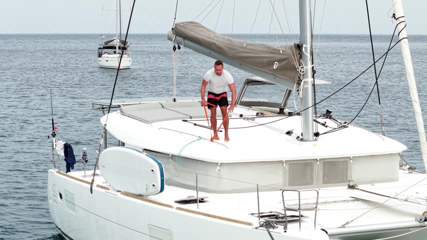 Morning on a yacht. Fitness exercises on the roof of a catamaran. A man doing exercises during a sea voyage. A man doing fitness exercises with a rubber elastic band