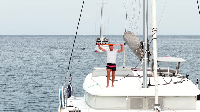 Morning on a yacht. Fitness exercises on the roof of a catamaran. A man doing exercises during a sea voyage. A man doing fitness exercises with a rubber elastic band
