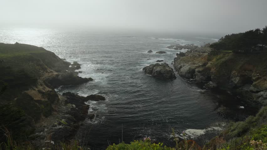 Rocky coastline with crashing waves and fog rolling in over the Pacific Ocean in Big Sur, California.
