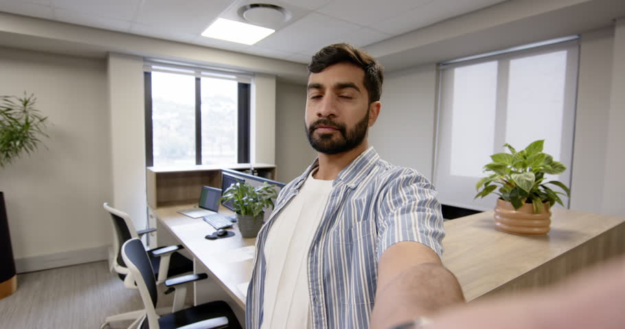 Pointing Indian man showing modular desks in modern coworking space with smartphone, copy space. Technology, workspace, modern, professional, urban, productivity, interior
