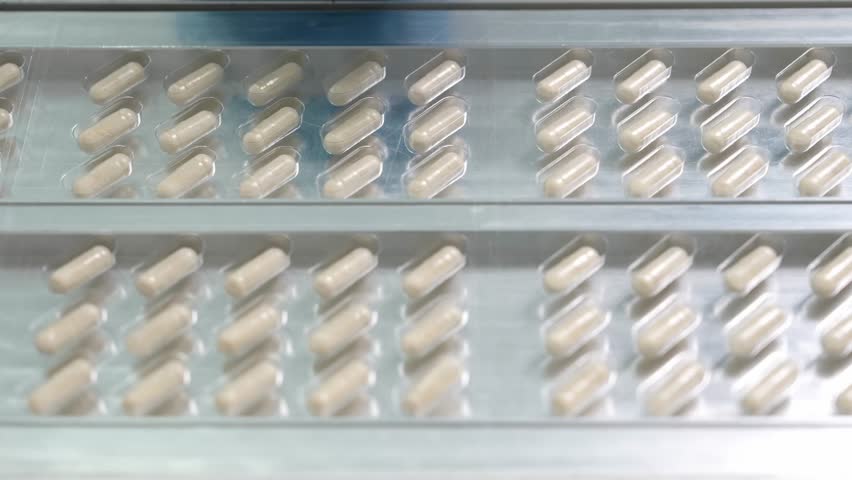 Capsules filled with medicine move along a pharmaceutical production line in transparent plastic blister packaging during the final packaging process inside a cleanroom facility.