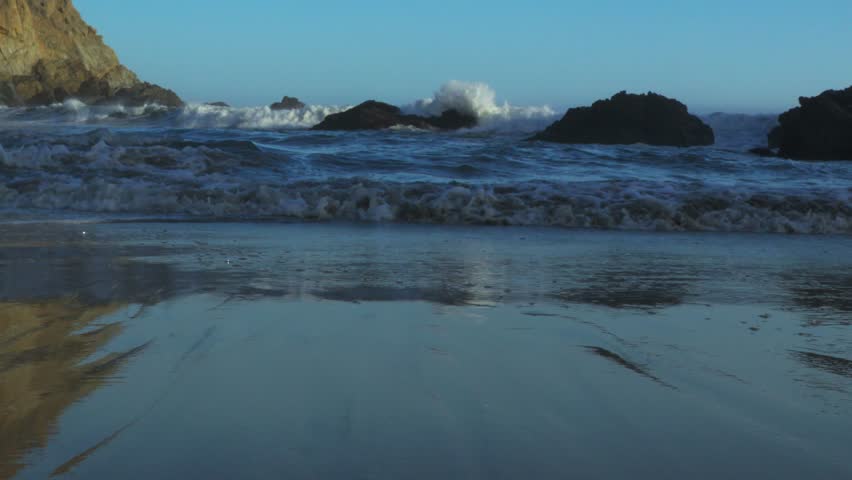 Ocean waves crashing against coastal rocks at Big Sur, California, with reflections of cliffs on the wet sandy beach under a clear blue sky.