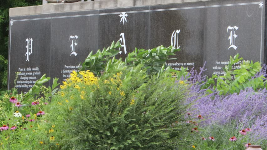 PEACE engraved on the side of a marble wall with flowers waving in the wind in front of it.