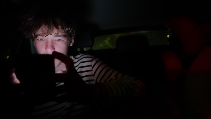 Teenage passenger immersed in smartphone screen while traveling inside darkened vehicle, illuminated by digital device glow during nighttime urban journey