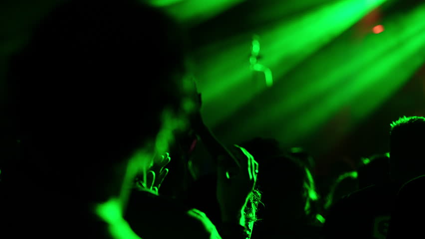 People are clapping and cheering during a live music concert, illuminated by green stage lights, creating a vibrant and energetic atmosphere