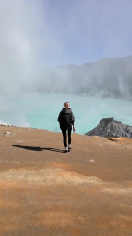 Aerial view of woman walking towards the edge of Ijen Volcano crater lake in East Java, Indonesia. Turquoise water, sulfur steam, and surreal volcanic landscape. Concept of nature, travel, adventure.