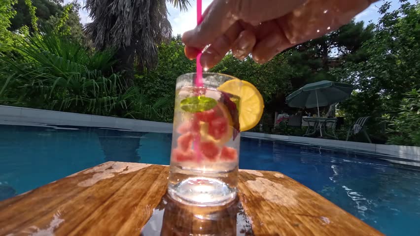 Hand stirring a cooling summer cocktail by the pool, relaxed vacation moment in sunny setting.