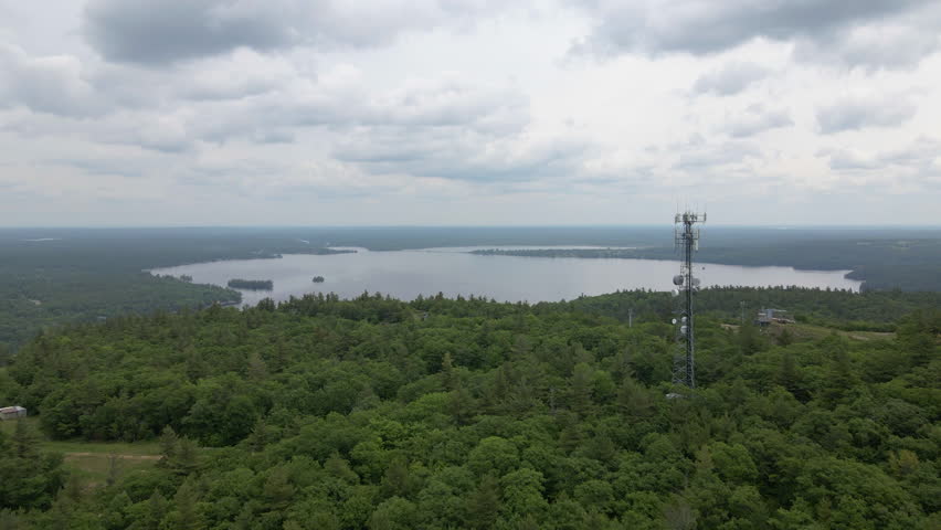 Aerial shot over green tree covered mountain past radio tower towards lake Calabogie on a cloudy day. Ontario Canada