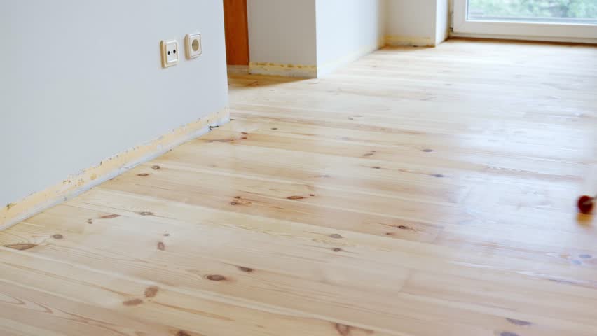A roller on an extension handle spreads transparent primer on a light pinewood floor in a modern indoor space with visible outlet and door frame, home improvement scene, static shot, real time
