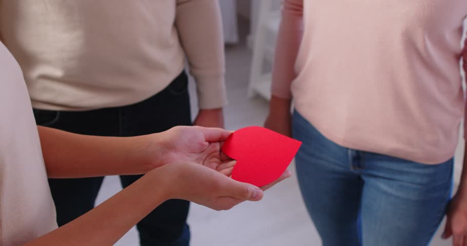 Closeup of family holding red paper heart in hands together standing at home. Parents with children sharing love promoting charity. Health care, insurance and organ donation concept. 4k video.