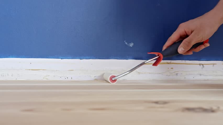 Person uses a detail roller to apply primer at the base of a blue wall above the skirting line while preparing for painting and floor treatment, static close-up, real time