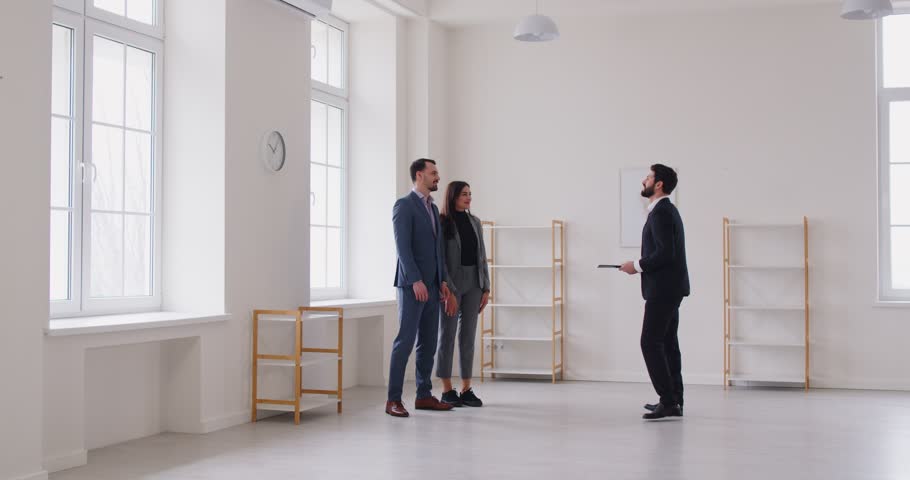 Happy smiling young couple customers shaking hands greeting professional man realtor. Male agent showing new apartments to clients discussing contract. Real estate market concept. 4k video.