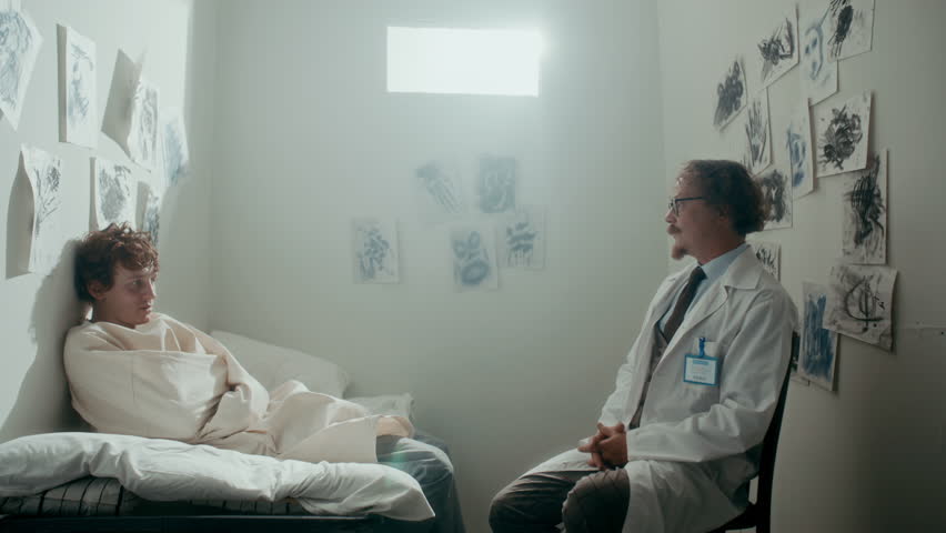 Psychiatrist talking to young male patient in straitjacket suffering from mental disorder as visiting him in seclusion room with abstract ink drawings on walls. Zoom shot