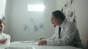 Professional doctor attentively listening to young male patient in straitjacket sitting across table in psychiatric ward with abstract drawings covering walls. Zoom-out shot - Powered by Shutterstock - Get 15% off with code: PIKWIZARD15