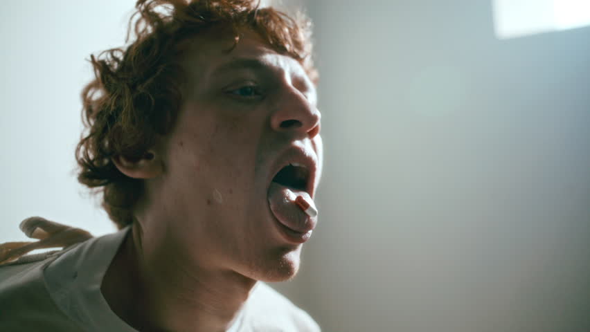 Young male patient in straitjacket showing pill on his tongue while taking medication under clinical supervision in psychiatric hospital