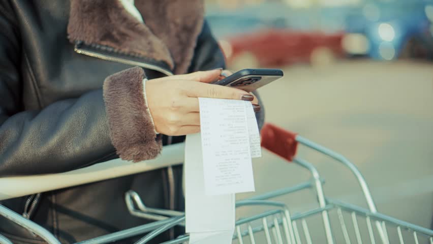 woman holds a long grocery receipt and her smartphone possibly checking expenses or using a budgeting app after shopping. Money management paying for product. Checking expenses smart spending shopping