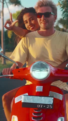 Young happy couple cruises on a red scooter during golden hour in Bali. The woman flashes a peace sign while enjoying the scenic views on a tropical vacation.