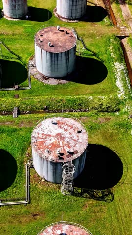 Rusty old oil tanks in overgrown green field in a neglected industrial compound, vertical drone video