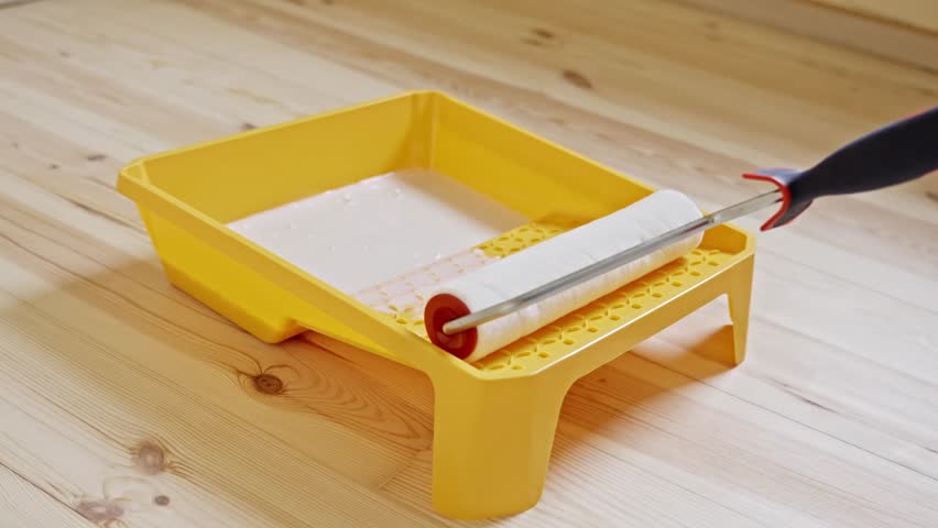 Close-up of a paint roller being loaded with wood primer from a yellow tray, preparing to coat a freshly sanded pine hardwood floor before varnish application, static camera, real-time