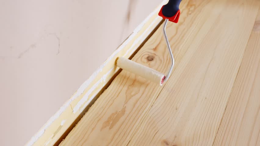 Close-up of hand using a narrow paint roller to apply primer along the baseboard edge of a softwood floor during a home renovation project, real time, static camera, early coating phase