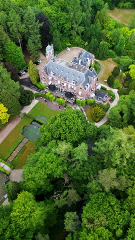 An aerial view of a historic mansion surrounded by lush green trees and a beautifully landscaped garden with a fountain, walking paths, and parking area. The scene shows vibrant summer greenery.